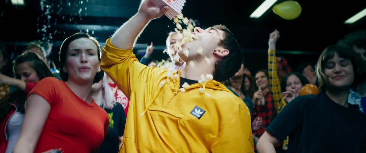 Standbild aus God Damn. Ein junger Mann im gelben Pullover schüttet sich auf einer Party Popcorn in den Mund, um ihn herum Feiernde.