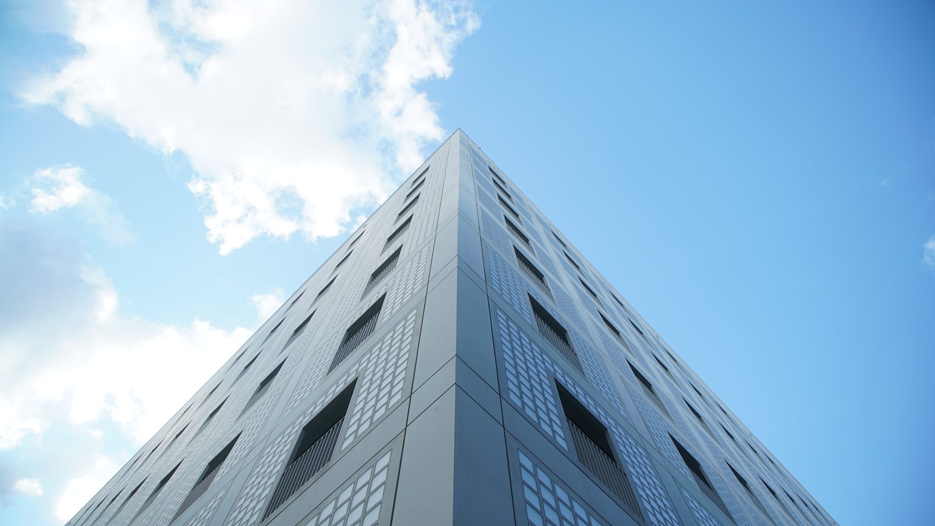 Gebäudeecke mit gerasterter Fassade von unten vor blauem Himmel mit Wolken.