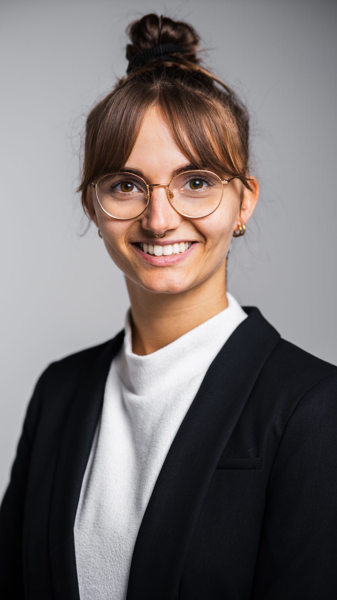 Studioporträt: lächelnde Frau mit runder Brille und Dutt in schwarzem Blazer vor grauem Hintergrund.