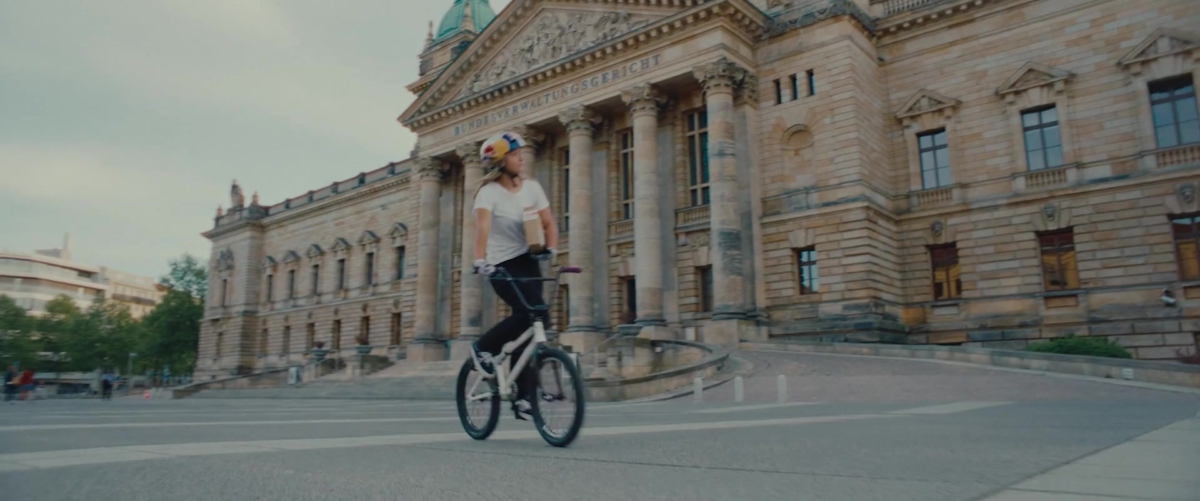 Standbild aus RB Leipzig – The Ultimate Jersey Journey. Eine BMX-Fahrerin mit Helm fährt mit einem Paket unter dem Arm vor einem historischen Gebäude.