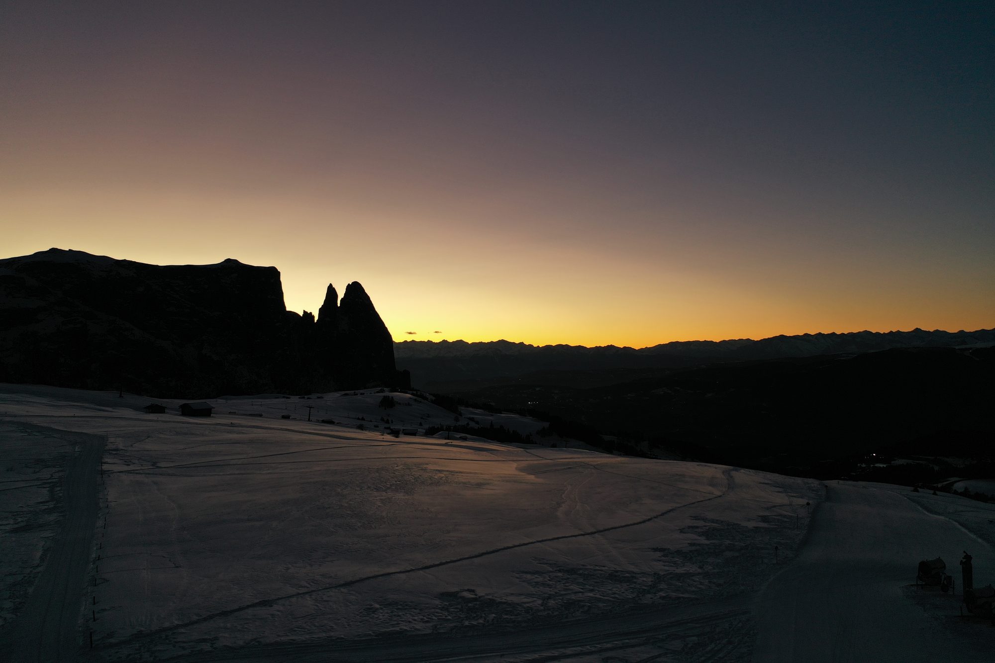 Bergsilhouette mit Felszacken vor orangefarbenem Horizont, davor verschneite Almwiesen in der Dämmerung.
